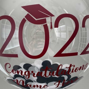 May include: A transparent balloon featuring the year "2022" and a burgundy graduation cap. The balloon has the words "Congratulations Name Here" in burgundy, and is filled with black, white, and grey confetti.
