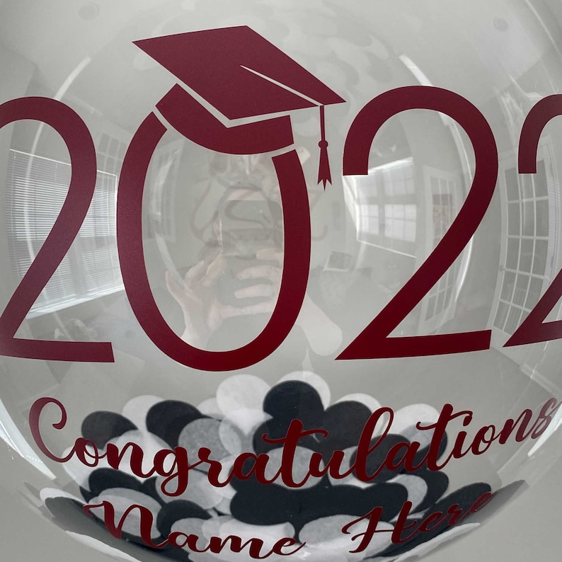 May include: A transparent balloon featuring the year "2022" and a burgundy graduation cap. The balloon has the words "Congratulations Name Here" in burgundy, and is filled with black, white, and grey confetti.