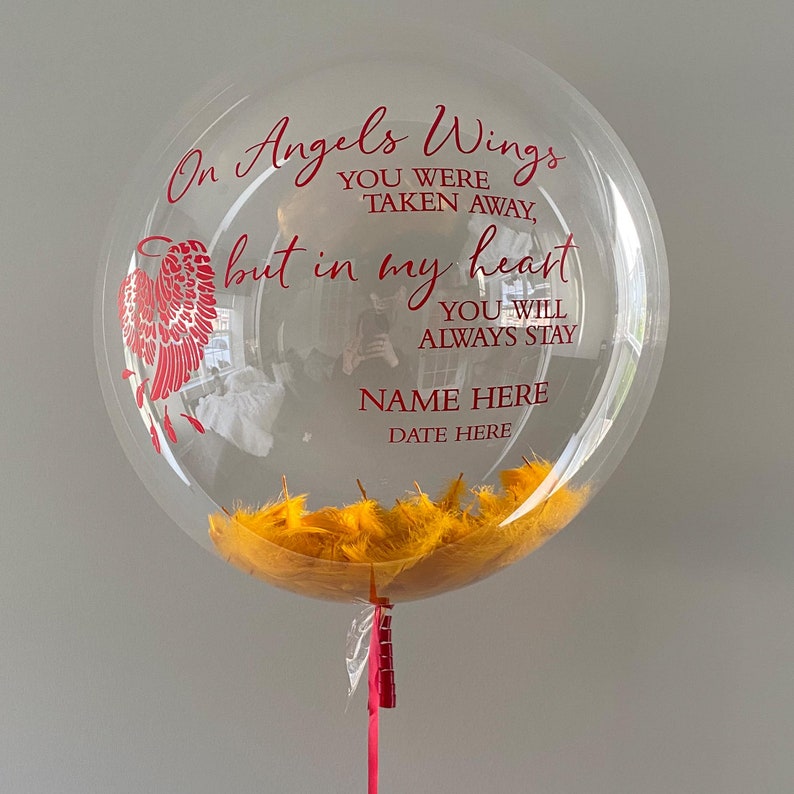 K&ouml;nnte beinhalten: Ein transparenter Ballon mit gelben Federn im Inneren. Der Ballon tr&auml;gt eine rote Aufschrift mit dem Text "On Angels Wings You Were Taken Away, But In My Heart You Will Always Stay. Name Here Date Here."