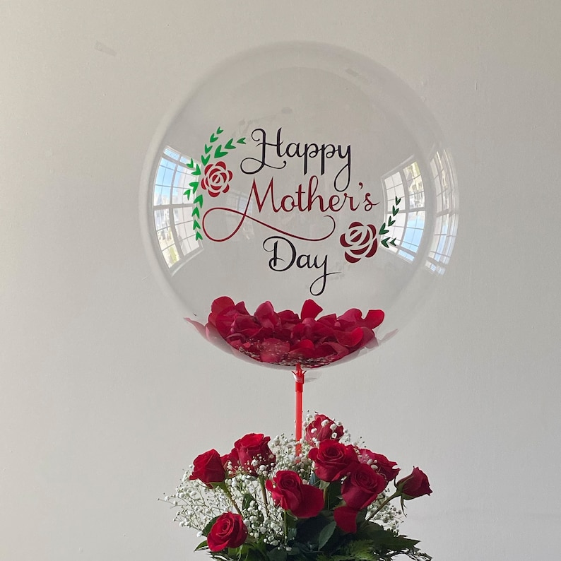 Peut inclure: Un ballon transparent rempli de p&eacute;tales de roses rouges avec le texte "Happy Mother's Day" en lettres vertes et rouges. Un bouquet de roses rouges avec de la gypsophile blanche est au premier plan.