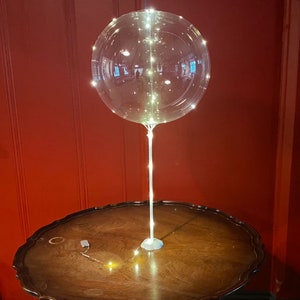 Puede incluir: Un globo transparente con luces LED blancas en su interior, suspendido de una cuerda blanca, sobre una mesa de madera.