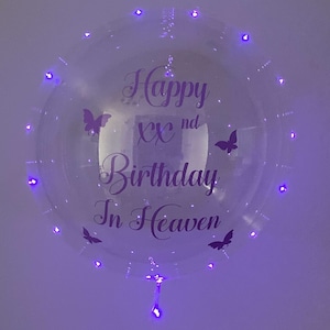 May include: A clear, round balloon with purple LED lights and the text "Happy XXnd Birthday In Heaven" in a decorative font. The balloon is adorned with butterfly silhouettes.