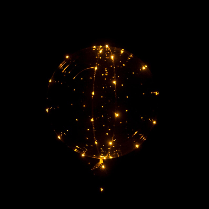Puede incluir: Un globo transparente con luces LED blancas c&aacute;lidas enroscadas a su alrededor. El globo est&aacute; inflado y parece flotar en una habitaci&oacute;n oscura.