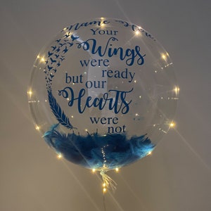 Peut inclure: Un ballon transparent avec des plumes bleues et des lumières blanches. Le ballon porte une inscription bleue qui dit "Tes ailes étaient prêtes mais nos cœurs ne l'étaient pas".