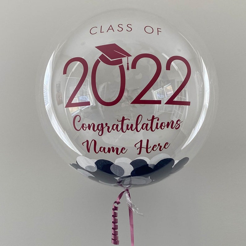 May include: A transparent graduation balloon with burgundy text reading "CLASS OF 2022", a mortarboard, and "Congratulations Name Here". The balloon contains confetti and is tied with a pink ribbon.