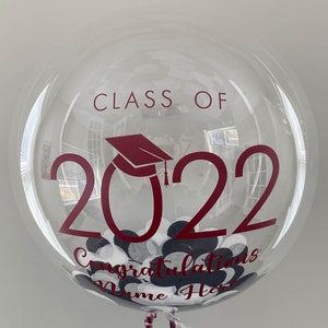 May include: A clear bubble balloon with maroon text reading "CLASS OF 2022" and "Congratulations Name Here". The balloon is filled with white, black, and dark maroon confetti. A graduation cap graphic is also visible.