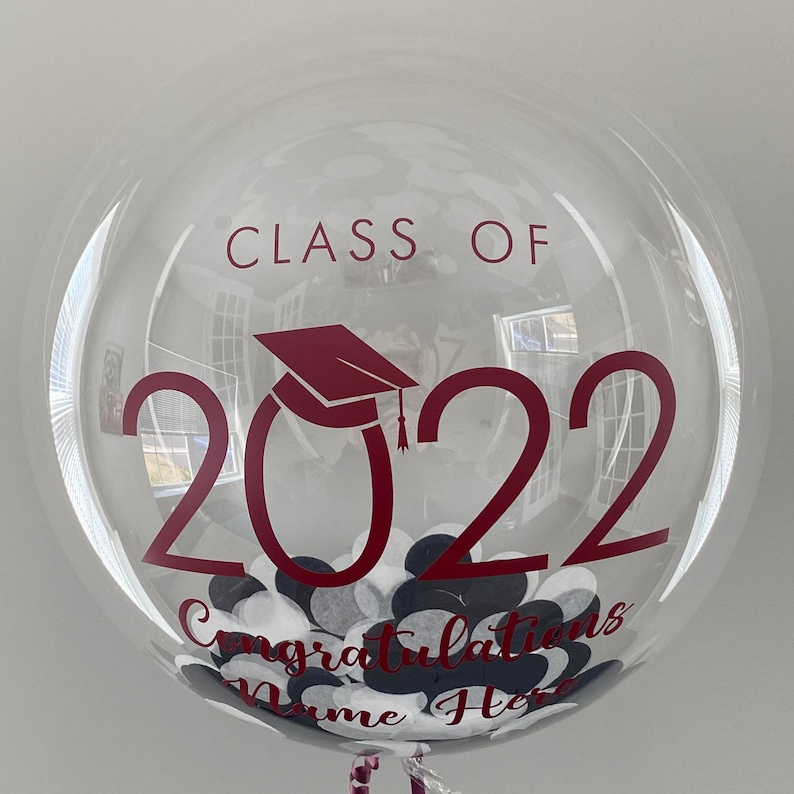 May include: A clear bubble balloon with maroon text reading "CLASS OF 2022" and "Congratulations Name Here". The balloon is filled with white, black, and dark maroon confetti. A graduation cap graphic is also visible.