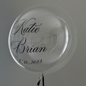 May include: A clear bubble balloon with black lettering that reads "Katie & Brian" and "Oct. 30, 2023".