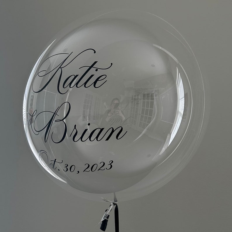 May include: A clear bubble balloon with black lettering that reads "Katie & Brian" and "Oct. 30, 2023".