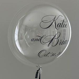 May include: A clear bubble balloon with black lettering that reads "Katie and Brian Oct. 30, 20..."