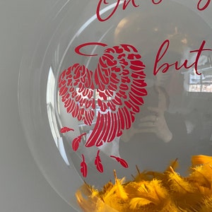 K&ouml;nnte beinhalten: Ein transparenter Glas-Anh&auml;nger mit einem roten herzf&ouml;rmigen Engelsfl&uuml;gel-Design und dem Text "On a wing but not forgotten". Der Anh&auml;nger ist mit gelben Federn gef&uuml;llt.