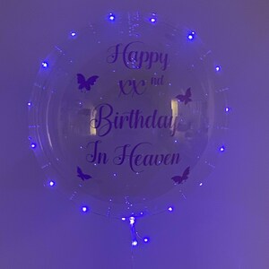 May include: A clear, round balloon with the text "Happy XXnd Birthday In Heaven" in purple script, adorned with butterfly decals. The balloon is illuminated by small, blue LED lights around its perimeter.