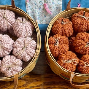 May include: Two round woven baskets with leather handles, each filled with multiple crocheted pumpkins. The left basket contains light pink pumpkins, while the right basket holds burnt orange pumpkins. Each pumpkin features a small brown stem.