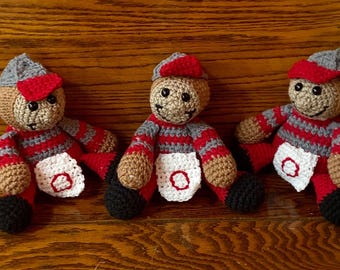 May include: Three handmade crochet dolls featuring tan faces, black eyes, and stitched smiles. Each doll wears a gray and red striped long-sleeve shirt, a gray cap with a red brim, black shoes, and a white apron with a red circular embroidered detail.