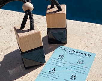 May include: Two square black glass car diffuser bottles with light wood caps and black hanging cords with wooden beads. A light blue instruction card is included with the text CAR DIFFUSER care instructions and various icons and safety warnings.
