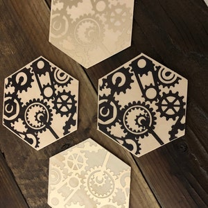 May include: Three hexagonal coasters with a gear pattern. The coasters are made of wood and have a light brown color. The gears are black and are arranged in a random pattern.