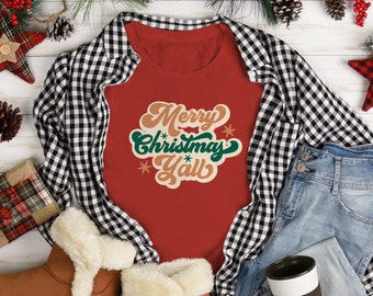 May include: A festive flat lay featuring a red t-shirt with "Merry Christmas Y'all" text, a black and white plaid shirt, blue jeans, brown boots with faux fur trim, and holiday decorations including pine cones and red ornaments.
