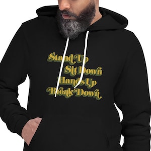 以下が含まれることがあります： 「Stand Up Sit Down Hands Up Black Down」と書いてある、金色と緑色のグラフィックが施された黒いパーカーです。