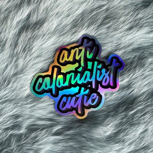 Könnte beinhalten: Ein holografischer Aufkleber mit dem Text "anti colonialist cutie" in einem Regenbogen-Farbschema.