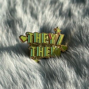 Puede incluir: Pin de esmalte dorado con las palabras "THEY/THEM" en una fuente retro con acentos amarillos, verdes y rosas. El pin tiene un pequeño corazón a cada lado y una estrella en la parte superior.