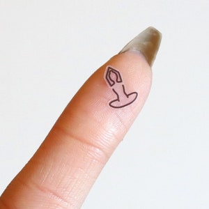 May include: A small, black line drawing of a person sitting in a yoga pose on a finger.