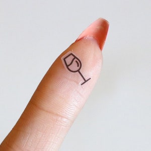 May include: A small, transparent sticker featuring a minimalist black outline of a wine glass. The sticker is placed on a fingertip against a white background, highlighting the simple design.