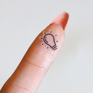 May include: A transparent lightbulb sticker with a black outline is placed on a finger. The sticker has a light pink hue and is positioned near the fingertip. The background is a plain white surface.