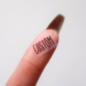 May include: A clear, rectangular sticker with the word "CUSTOM" printed in black cursive script. The sticker is placed on a finger, with a blurred background.