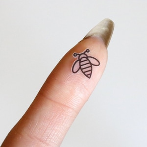Op de afbeelding: Een vinger met een kleine bij-sticker erop. De bij is zwart en heeft een lichtbruine streep.