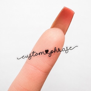 May include: A small, clear adhesive sticker featuring the black cursive text custom phrase with a small heart icon replacing the letter a. The sticker is thin and elongated, designed for application on a smooth surface or skin.