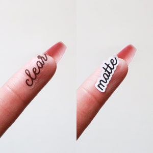 May include: Two fingers with clear and matte stickers on them. The stickers say "clear" and "matte" in cursive.