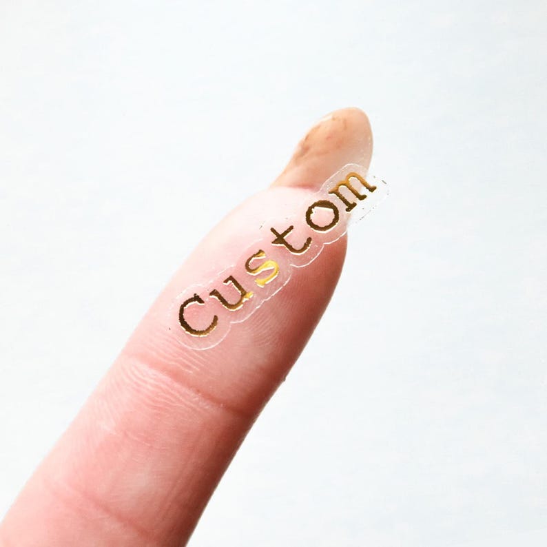 May include: A clear sticker with the word "Custom" in gold lettering. The sticker is on a finger, with a white background.