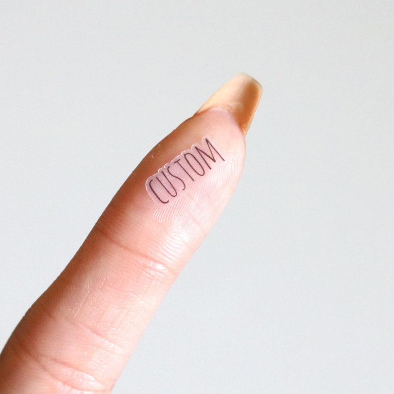 Puede incluir: Un dedo con una pegatina transparente que dice "CUSTOM" en ella.