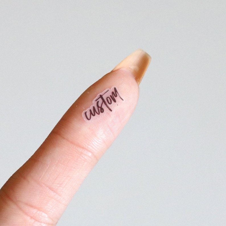 May include: A clear, rectangular sticker with the word "custom" in a handwritten font. The sticker is placed on a fingertip against a soft, neutral background. The nail is long and has a light, natural color.