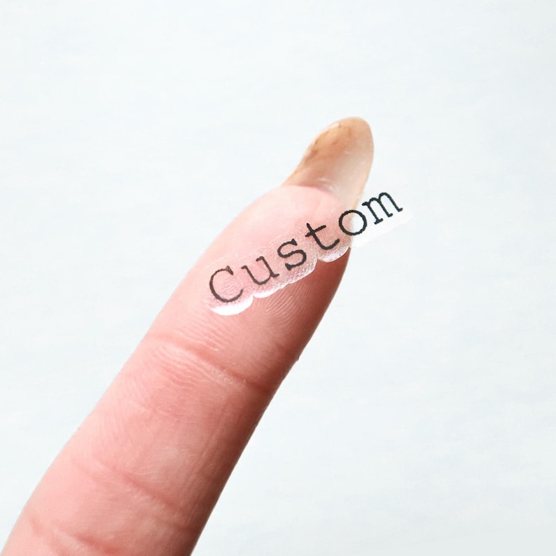 May include: A clear, scalloped-edge sticker with the word "Custom" printed in black. The sticker is held on a finger against a white background. The sticker is likely for personalized products.