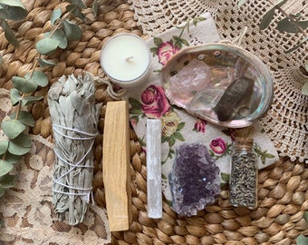 Peut inclure: Un ensemble d'objets pour un rituel spirituel, comprenant un bâtonnet de sauge blanche, du bois de palo santo, une bougie blanche, un petit pot de lavande séchée, un cristal de quartz clair, un cristal d'améthyste violet et un cristal de rose quartz rose dans une coquille de coquillage.