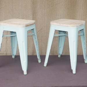 May include: Two light blue metal stools with square wooden seats. The stools have a distressed finish and are set against a neutral background. Each stool is approximately 18 inches tall.