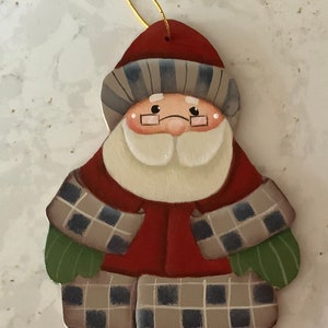 May include: A hand-painted wooden ornament of Santa Claus wearing a red coat, plaid scarf, and green mittens. He has a white beard and a red hat with a white trim.