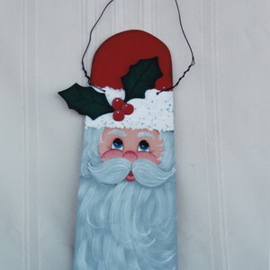 May include: A painted wooden Christmas ornament depicting Santa Claus with a white beard, red hat, and green holly. The ornament has a wire hanger for hanging.