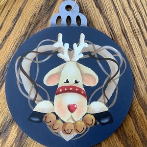 May include: A navy blue Christmas ornament featuring a reindeer design. The reindeer has a red collar, a heart-shaped nose, and is surrounded by a wreath. Gold-colored bells hang below the reindeer. The ornament has a silver top and a gold string.