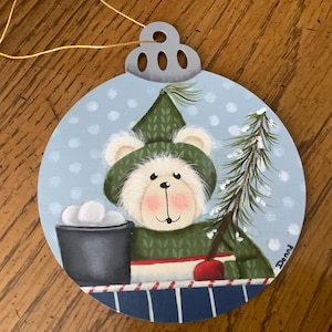 May include: A hand-painted wooden ornament featuring a white bear wearing a green hat and sweater, holding a bucket of snowballs. The bear is standing in front of a small pine tree with snow on the branches. The ornament is painted on a blue background with white dots and has a silver hanger.