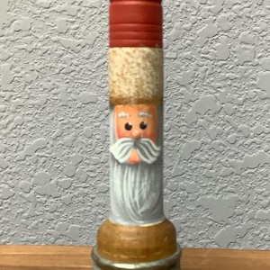 May include: A decorative bottle stopper featuring a Santa Claus design. The top is red, followed by a tan section, a painted face with a white beard, and a brown base with silver rings. A festive item for Christmas.