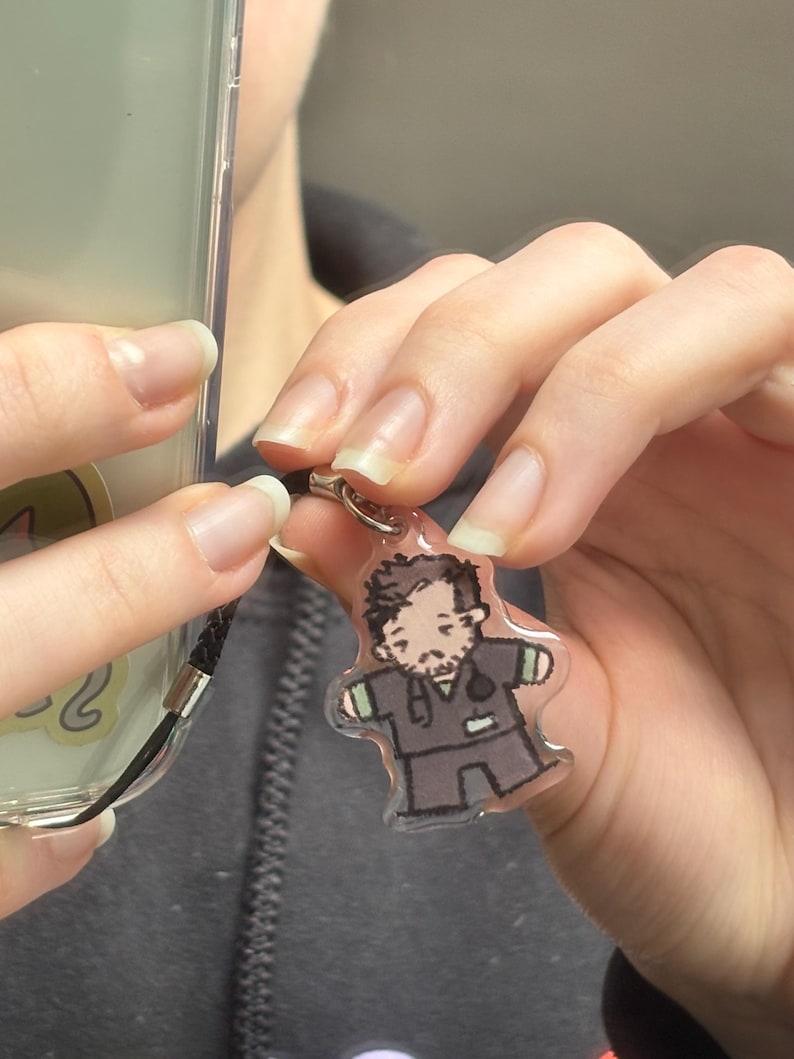 May include: A clear acrylic charm featuring a cartoon illustration of a person with brown hair and a beard, wearing a dark gray scrub top with green sleeve cuffs and a stethoscope around their neck, attached to a black cord phone strap.
