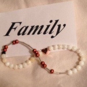 Puede incluir: Dos pulseras de cuentas en forma de símbolo de infinito, con una tarjeta que dice "Family". Una pulsera tiene cuentas blancas y la otra cuentas blancas y rojas. Un pequeño dije de corazón está entre las pulseras. El fondo es de tela beige claro.