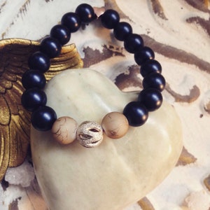 Könnte beinhalten: Ein Armband mit mattschwarzen Perlen und drei dekorativen Perlen. Die zentrale Perle ist silberfarben und strukturiert, flankiert von zwei beigefarbenen Perlen mit einem Muster. Das Armband wird auf einem herzförmigen Objekt präsentiert.
