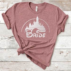 Puede incluir: Una camiseta color malva con un gráfico blanco de un castillo y la palabra "BRIDE" en una fuente estilizada.