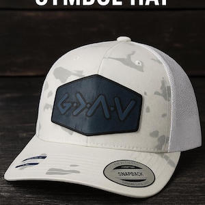 May include: White and gray camouflage-patterned snapback hat with a navy blue patch featuring the text "G>A>V". The hat has a white mesh back and a curved brim. The text "GOD IS GREATER SYMBOL HAT" is at the top.