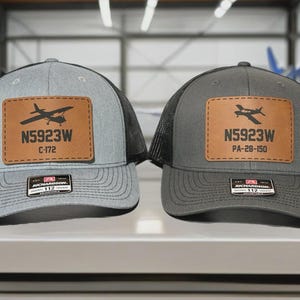 May include: Two trucker hats with a patch featuring an airplane silhouette and the text "N5923W". One hat is gray with a black mesh back, the other is dark gray with a black mesh back. The patches are brown.