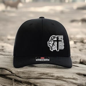May include: Black baseball cap with a curved brim. The cap features a white graphic of a lion's head, a cross, and the words "MAN OF GOD HUSBAND DAD HERO". The cap is sitting on a weathered wooden surface.