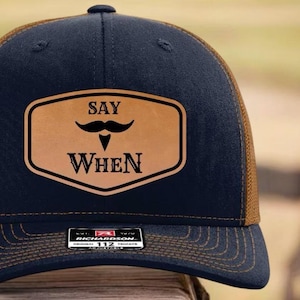 May include: Navy blue trucker hat with a brown mesh back and brim. The front features a brown leather patch with a black mustache graphic and the words "SAY WHEN". A small tag is visible on the brim.